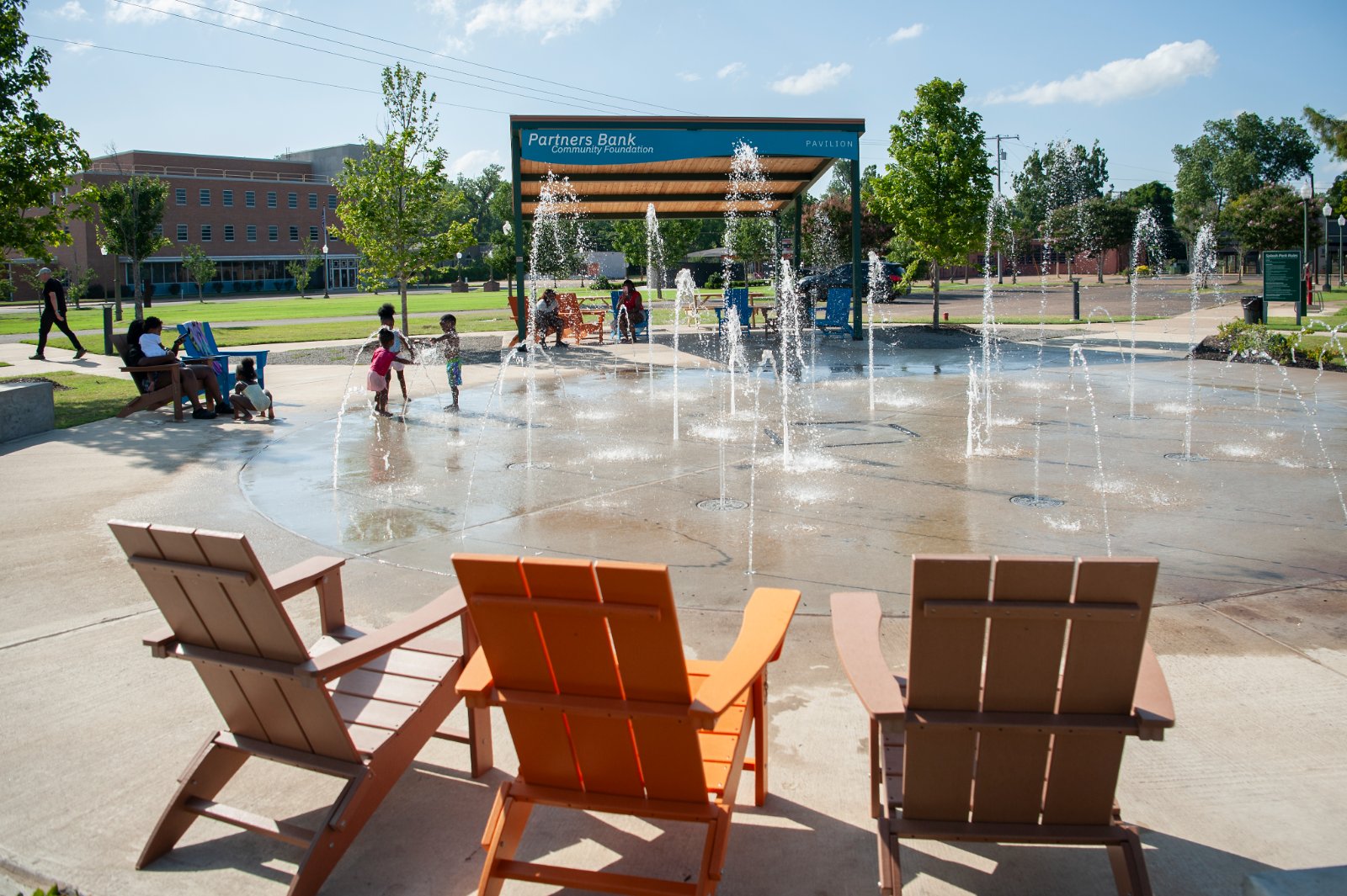 Children playing in the splash pad jets with bright Adirondack chairs in the foreground and the Partners Bank Pavilion beyond
